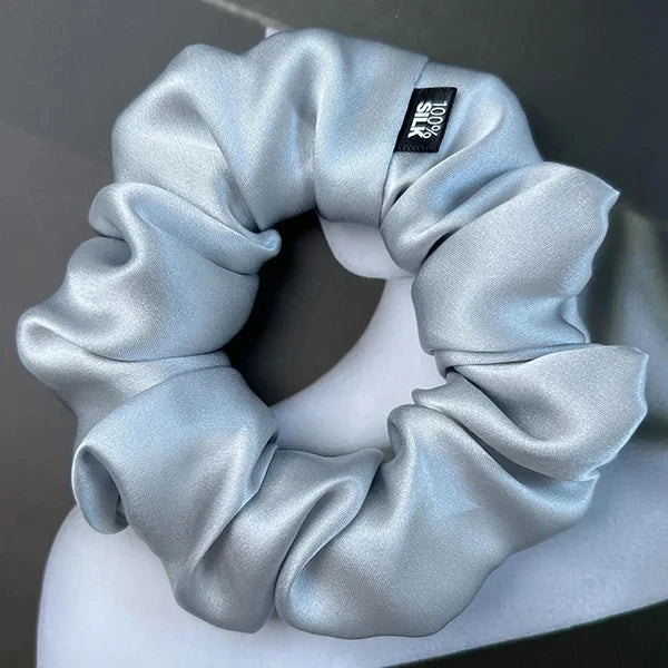 Silver silk scrunchie displayed on a white stand against a gray background.