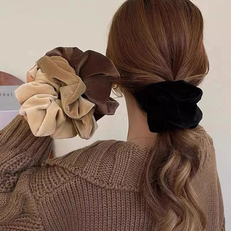 Model wearing a black velvet scrunchie in a low ponytail, holding beige, coffee and brown scrunchies beside her hair.