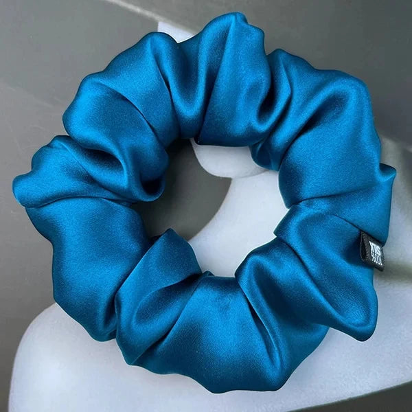 Peacock blue silk scrunchie displayed on a white stand against a gray background.