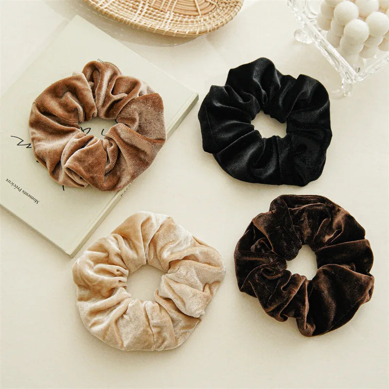 Four velvet scrunchies in coffee, black, beige and dark brown laid flat on a cream surface.