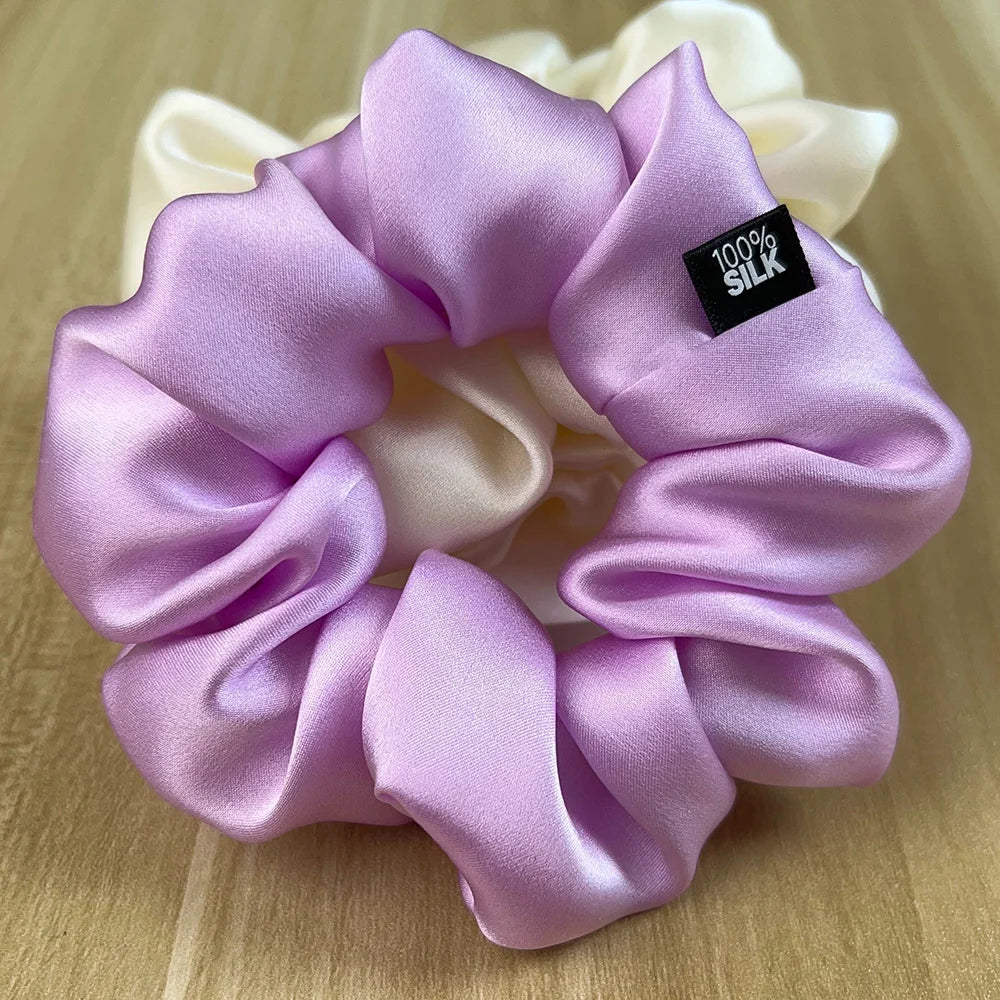 Close-up of a lavender silk scrunchie with a black label, resting in front of an ivory scrunchie.